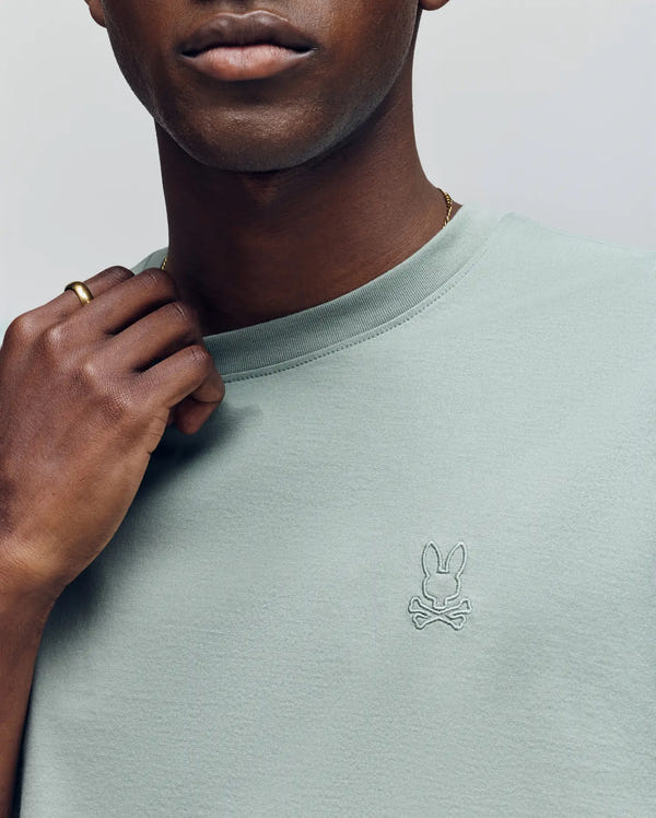 A man wears the Psycho Bunny MENS JAMES PREMIUM CREW NECK TEE (B6U952D200) in light green, crafted from soft Pima cotton with a small embroidered bunny and crossbones logo, holding his collar and wearing a gold ring.
