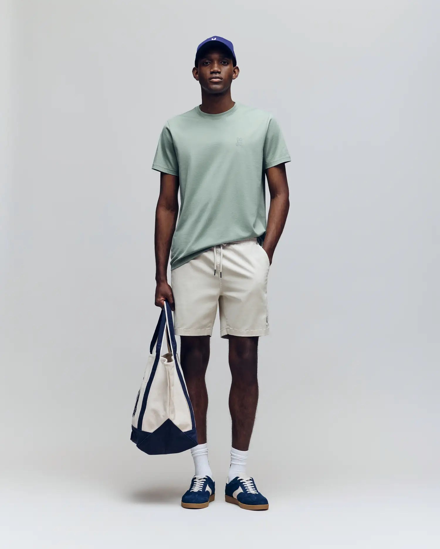 A man stands against a light background wearing the Psycho Bunny MENS JAMES PREMIUM CREW NECK TEE (B6U952D200) in light green, paired with beige shorts, white socks, blue sneakers, a blue cap, and holding a beige and navy tote bag.