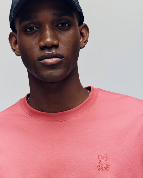 A man wearing a black cap and Psycho Bunny's MENS JAMES PREMIUM CREW NECK TEE in pink Pima cotton, featuring a small embroidered bunny and crossbones logo on the chest, looks at the camera with a neutral expression.