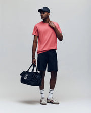 A man wearing a black cap, Psycho Bunny MENS JAMES PREMIUM CREW NECK TEE - B6U952D200 in pink, black shorts, striped white socks, and sneakers stands holding a black duffel bag with a white graphic against a plain background.