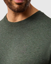 A close-up image shows a man wearing a forest green MENS JAMES PREMIUM CREW NECK TEE - B6U952D200 from Psycho Bunny. The stylish T-shirt is made from premium Pima cotton and features a small embroidered logo on the left side of the chest, showcasing the brand's signature stylized rabbit head and crossbones design. The man's lower face and part of his neck are visible.