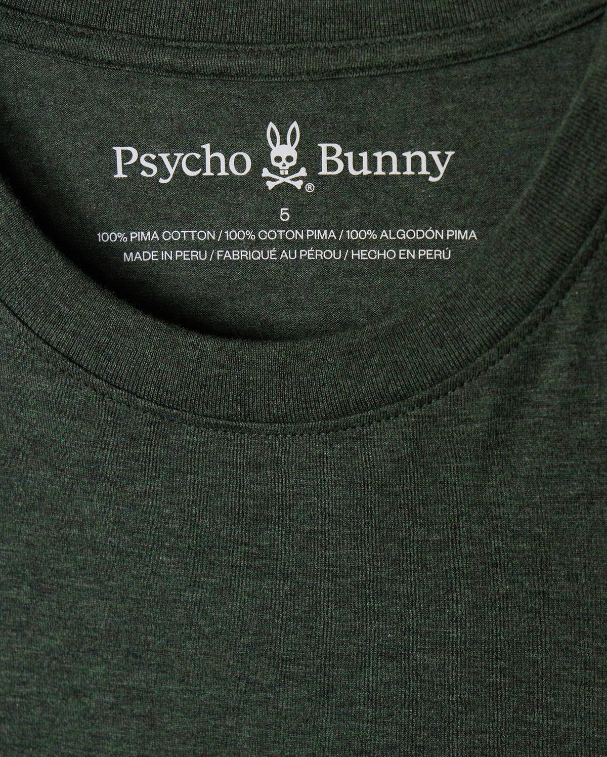 A close-up of the inside label of a dark green MENS JAMES PREMIUM CREW NECK TEE - B6U952D200, featuring the brand name "Psycho Bunny" along with a rabbit logo. The text indicates that the stylish crew neck shirt is made from 100% premium Pima cotton and was made in Peru.