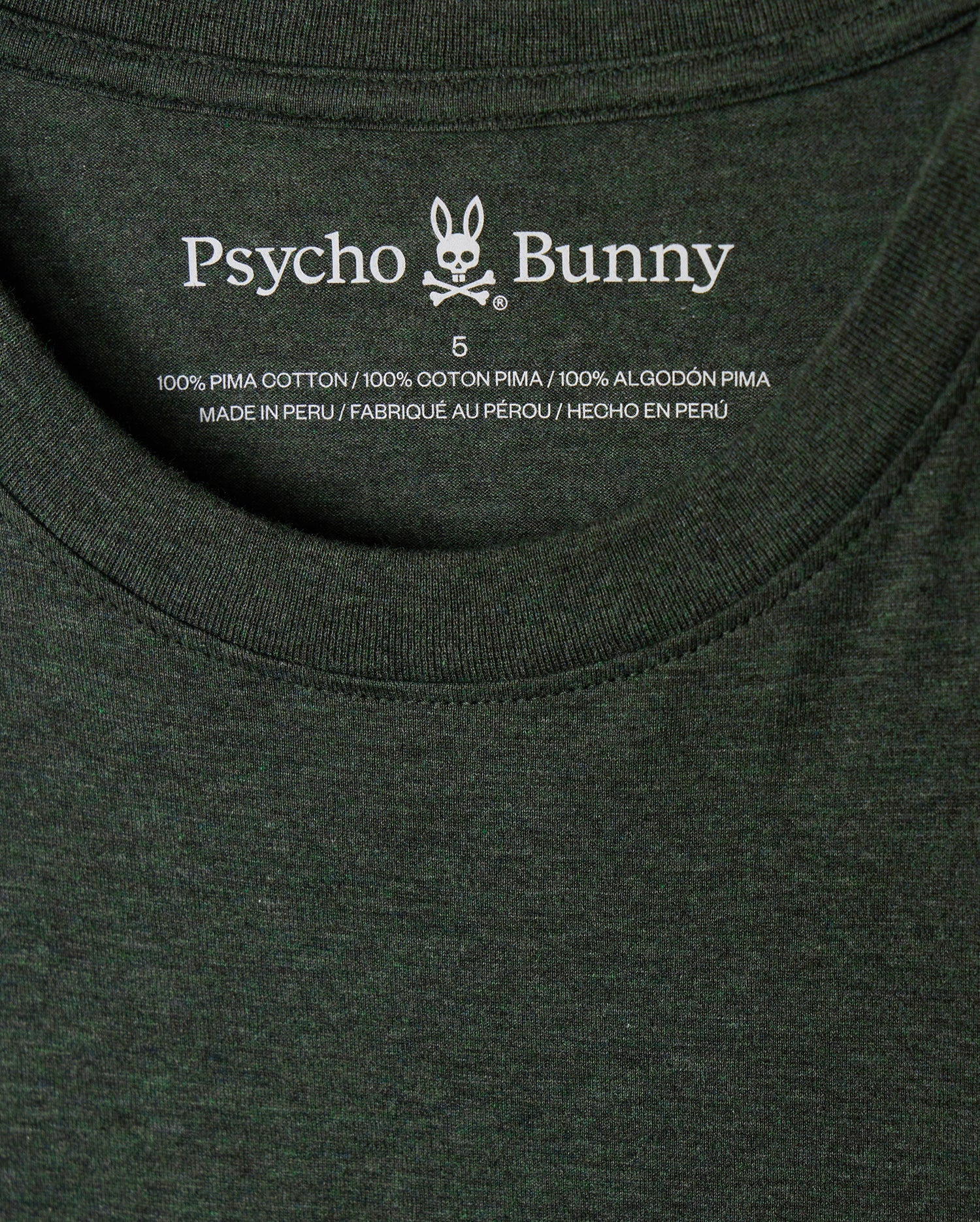 A close-up of the inside label of a dark green MENS JAMES PREMIUM CREW NECK TEE - B6U952D200, featuring the brand name "Psycho Bunny" along with a rabbit logo. The text indicates that the stylish crew neck shirt is made from 100% premium Pima cotton and was made in Peru.