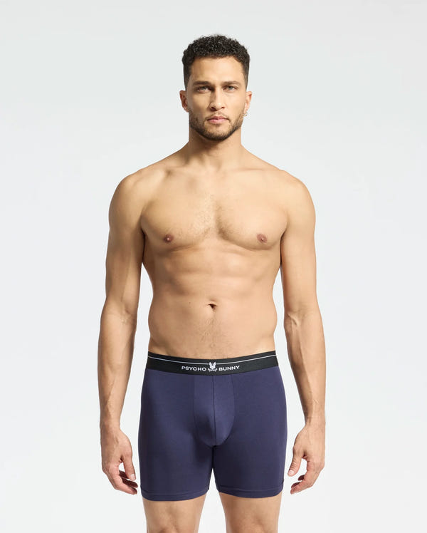 A man with short dark hair and a neutral expression models the Psycho Bunny MENS 2 PACK BOXER BRIEF - B6V481F200 against a plain background, showcasing navy blue Pima cotton with a black waistband featuring white text.