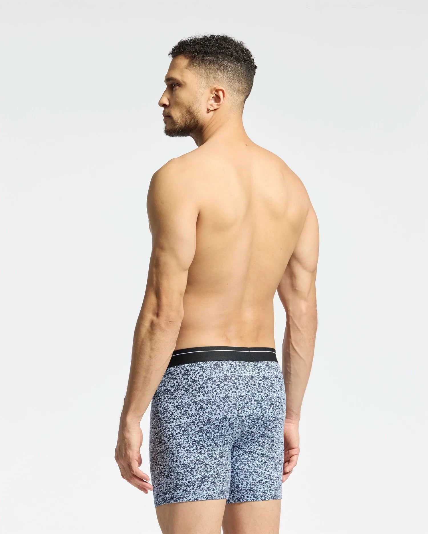 A man, shirtless in a white studio, glances over his shoulder wearing Psycho Bunny's MENS 2 PACK BOXER BRIEF - B6V481F200. Featuring a black waistband and crafted from a Pima cotton-Modal blend for all-day comfort.