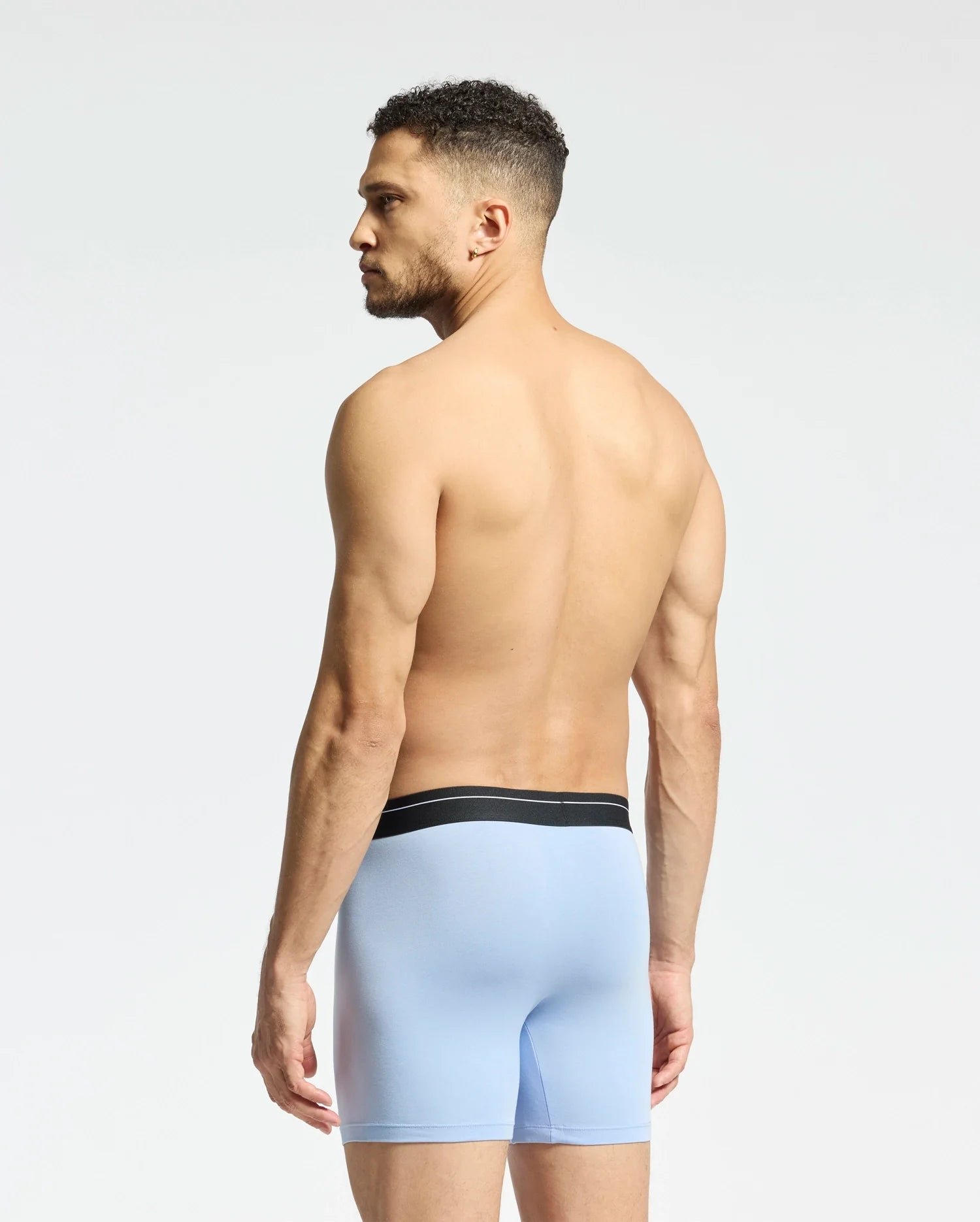 A man with short curly hair faces away from the camera, shirtless and gazing sideways against a neutral backdrop, wearing Psycho Bunny's MENS 2 PACK BOXER BRIEF - B6V481F200 in light blue modal blend featuring a black waistband.