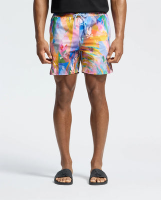 Wearing Psycho Bunny's MENS MILOS PRINTED SWIM TRUNK - B6W446F200 in vibrant geometric patterns of orange, purple, and blue, and black logo slides, a person stands against a white backdrop with hands relaxed by their sides.