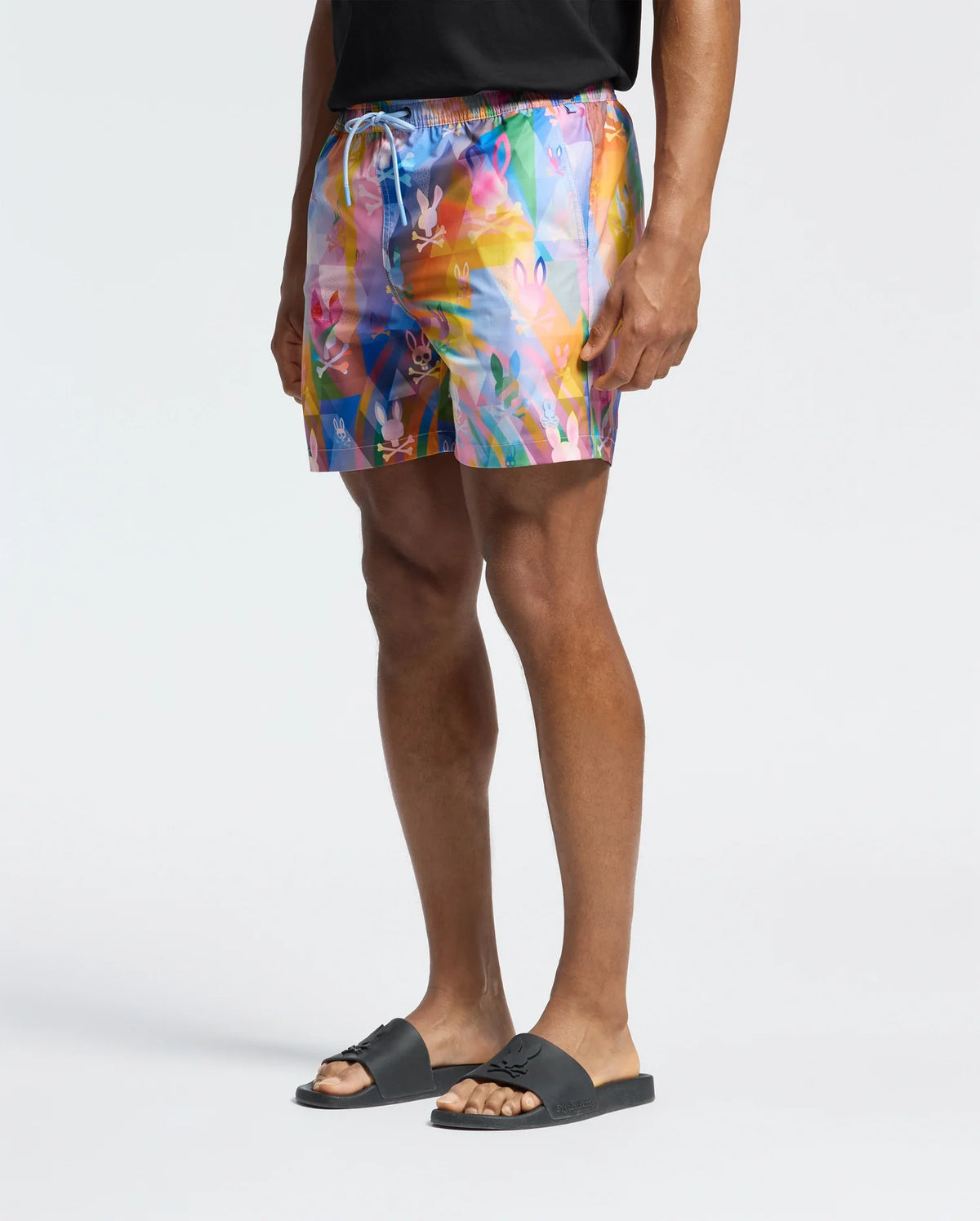 A person showcases vacation style in Psycho Bunny MENS MILOS PRINTED SWIM TRUNK with an abstract design, paired with a black shirt and slide sandals, set against a white background.