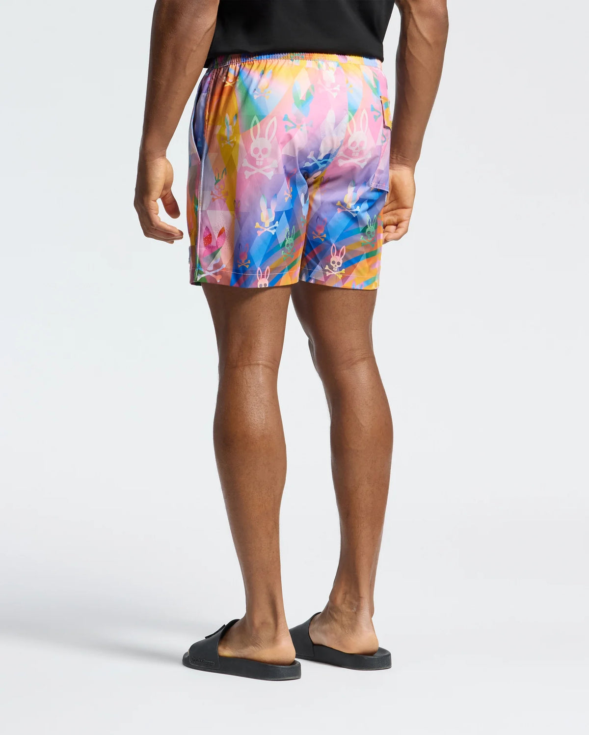A person is standing, facing away, in Psycho Bunny's colorful MENS MILOS PRINTED SWIM TRUNK - B6W446F200 with an abstract pattern featuring small white symbols, paired with a black T-shirt and slides. The plain light gray background perfectly showcases their vacation style.