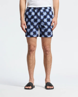 A person is sporting Psycho Bunny's MENS PAROS PRINTED SWIM TRUNK - B6W450F200, featuring a blue and black geo-pattern with a white waistband string. They're in black slide sandals, a partially visible white top, all set against a plain white background.