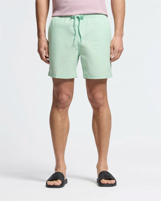 A person in mint green MENS SAMOS HYDROCHROMIC PRINTED SWIM TRUNK - B6W453F200 by Psycho Bunny and black slides stands facing forward, partially visible light pink shirt with a playful bunny pattern. The background is plain white.