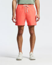A person is wearing Psycho Bunny's MENS SAMOS HYDROCHROMIC PRINTED SWIM TRUNK in coral pink with a white drawstring, paired with a green shirt and white sneakers, ready for the pool party against a crisp white background.