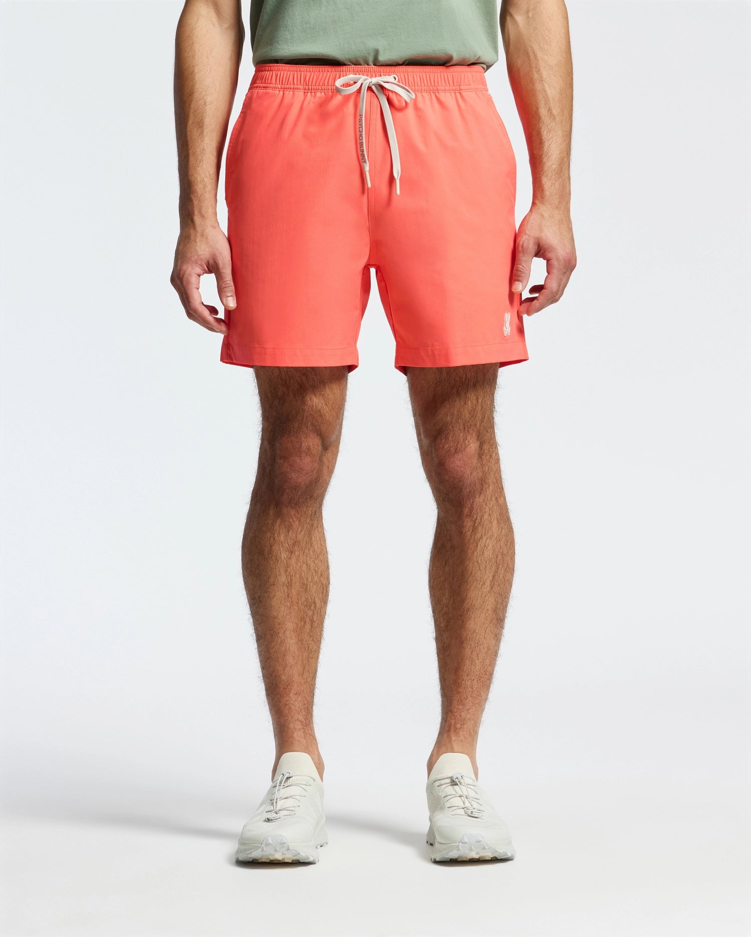 A person is wearing Psycho Bunny's MENS SAMOS HYDROCHROMIC PRINTED SWIM TRUNK in coral pink with a white drawstring, paired with a green shirt and white sneakers, ready for the pool party against a crisp white background.