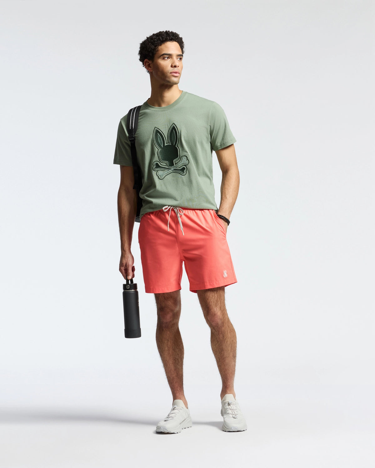 Against a plain backdrop, a person wears Psycho Bunny's green T-shirt with a bunny graphic and MENS SAMOS Hydrochromic Printed Swim Trunk in coral. They sport white sneakers, hold a hydrochromic black water bottle, have a bag strap over one shoulder, and gaze to the side.