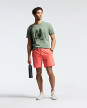 Against a plain backdrop, a person wears Psycho Bunny's green T-shirt with a bunny graphic and MENS SAMOS Hydrochromic Printed Swim Trunk in coral. They sport white sneakers, hold a hydrochromic black water bottle, have a bag strap over one shoulder, and gaze to the side.