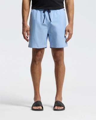 A person models Psycho Bunny's MENS SAMOS HYDROCHROMIC PRINTED SWIM TRUNK (B6W453F200) with an adjustable drawstring. Paired with a black shirt and slide sandals, only the lower body is shown against a white background.