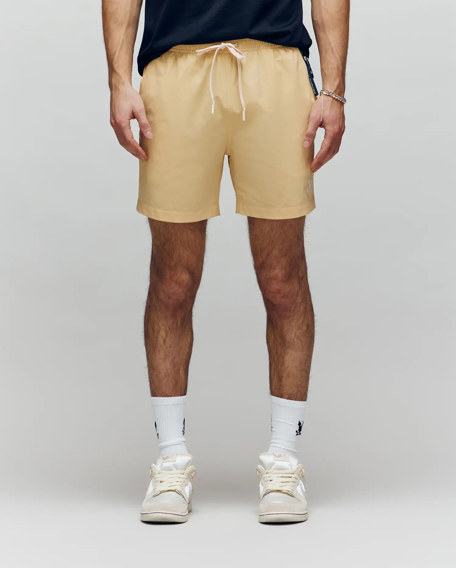 A person wearing Psycho Bunny MENS SAMOS HYDROCHROMIC PRINTED SWIM TRUNK (B6W453F200) in beige, paired with white crew socks and sneakers, stands against a plain light background. Only their lower torso and legs are visible.