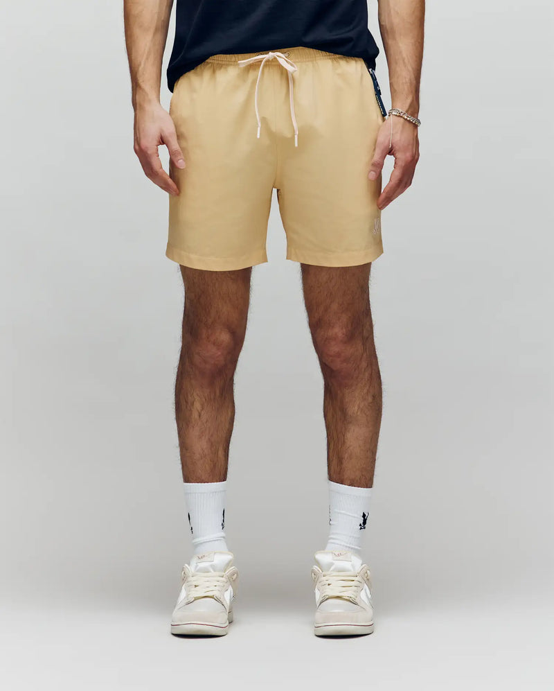 A person wearing Psycho Bunny MENS SAMOS HYDROCHROMIC PRINTED SWIM TRUNK (B6W453F200) in beige, paired with white crew socks and sneakers, stands against a plain light background. Only their lower torso and legs are visible.