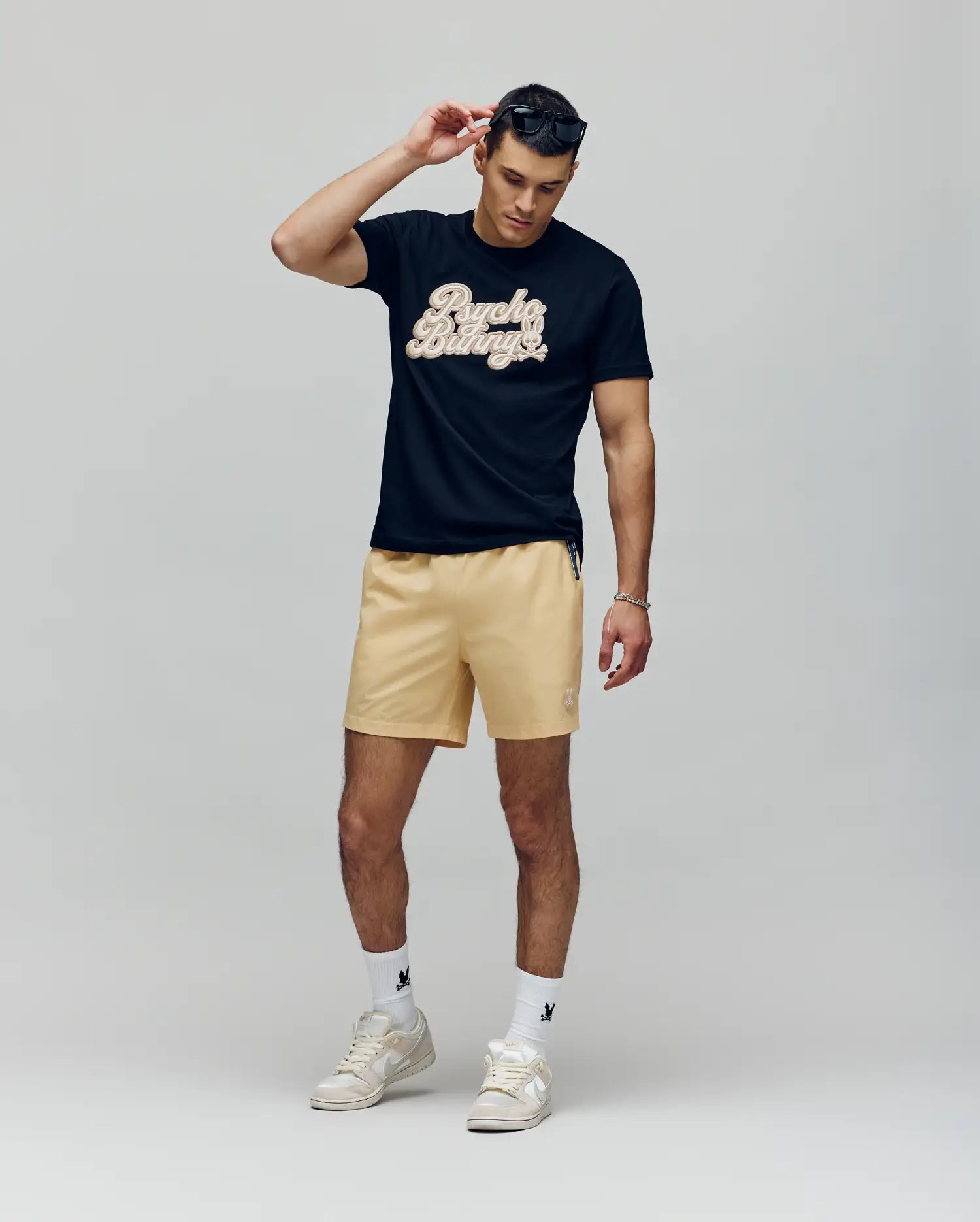 A man wearing a black graphic t-shirt, Psycho Bunny MENS SAMOS HYDROCHROMIC PRINTED SWIM TRUNK, white socks, and white sneakers adjusts his sunglasses with one hand while looking down against a plain background.