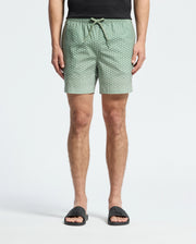 A person stands against a light background wearing Psycho Bunny's MENS BENDIGO ALL OVER PRINTED SWIM TRUNK, quick-dry green with white dots, paired with a black shirt and black slide sandals, showcasing effortless style.