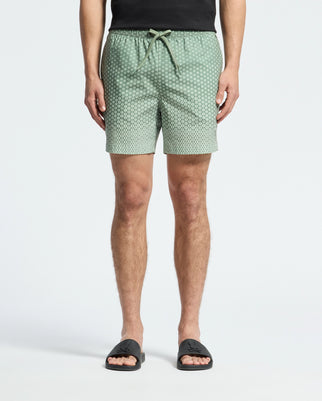 A person stands against a light background wearing Psycho Bunny's MENS BENDIGO ALL OVER PRINTED SWIM TRUNK, quick-dry green with white dots, paired with a black shirt and black slide sandals, showcasing effortless style.