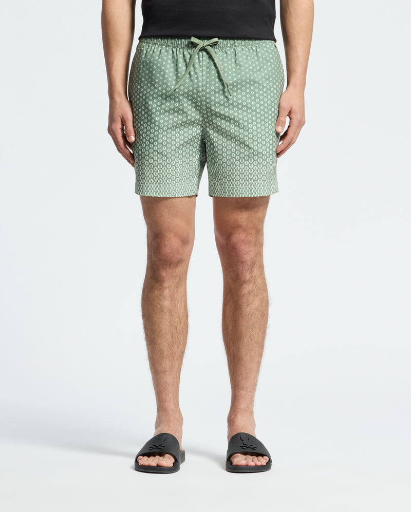 A person stands against a light background wearing Psycho Bunny's MENS BENDIGO ALL OVER PRINTED SWIM TRUNK, quick-dry green with white dots, paired with a black shirt and black slide sandals, showcasing effortless style.