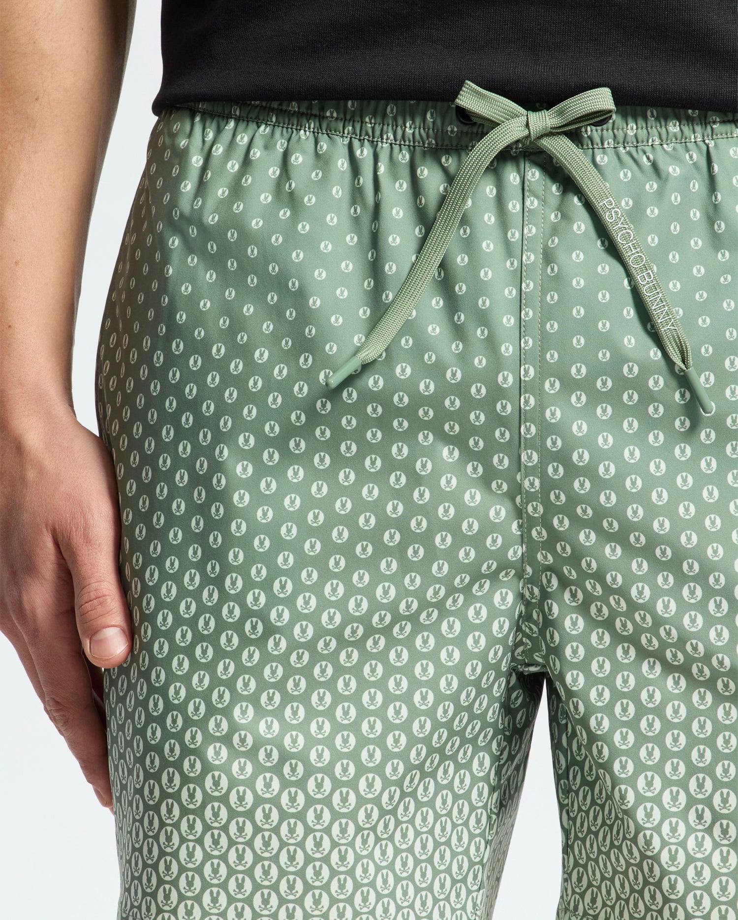 A person wears Psycho Bunny's MENS BENDIGO ALL OVER PRINTED SWIM TRUNK - B6W619F200, featuring a green leaf pattern and drawstring waistband. Their hand rests by their side, paired with a black top against a light background.