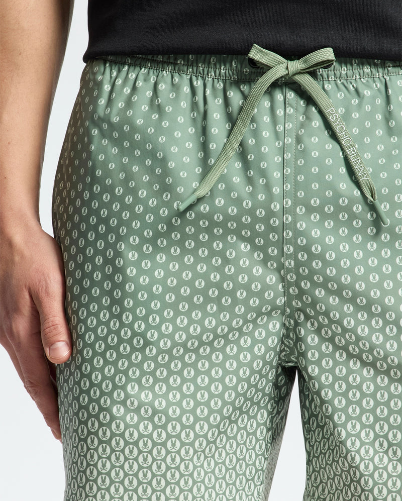 A person wears Psycho Bunny's MENS BENDIGO ALL OVER PRINTED SWIM TRUNK - B6W619F200, featuring a green leaf pattern and drawstring waistband. Their hand rests by their side, paired with a black top against a light background.