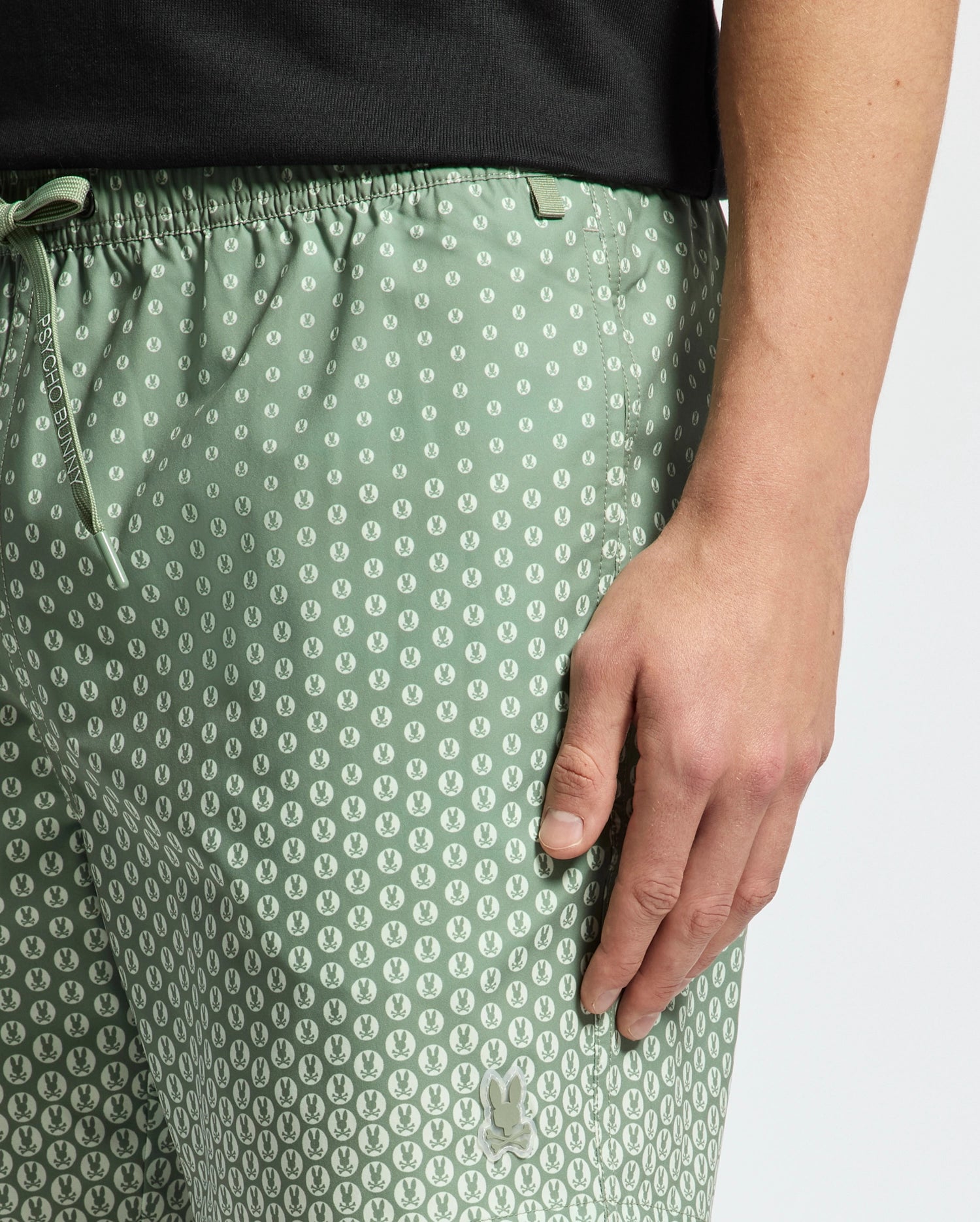 A person is pictured wearing the Psycho Bunny MENS BENDIGO ALL OVER PRINTED SWIM TRUNK, featuring a green pattern with small white dots and a drawstring. They pair it with a black top against a plain white background, resting their hand on their thigh.