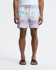 A person wears Psycho Bunny's MENS FORSTER BUNNY PRINTED SWIM TRUNK, a pastel-colored short with stars and bunnies featuring a pink drawstring, paired with a white shirt and black slide sandals against a plain white background. Their hands rest at their sides.