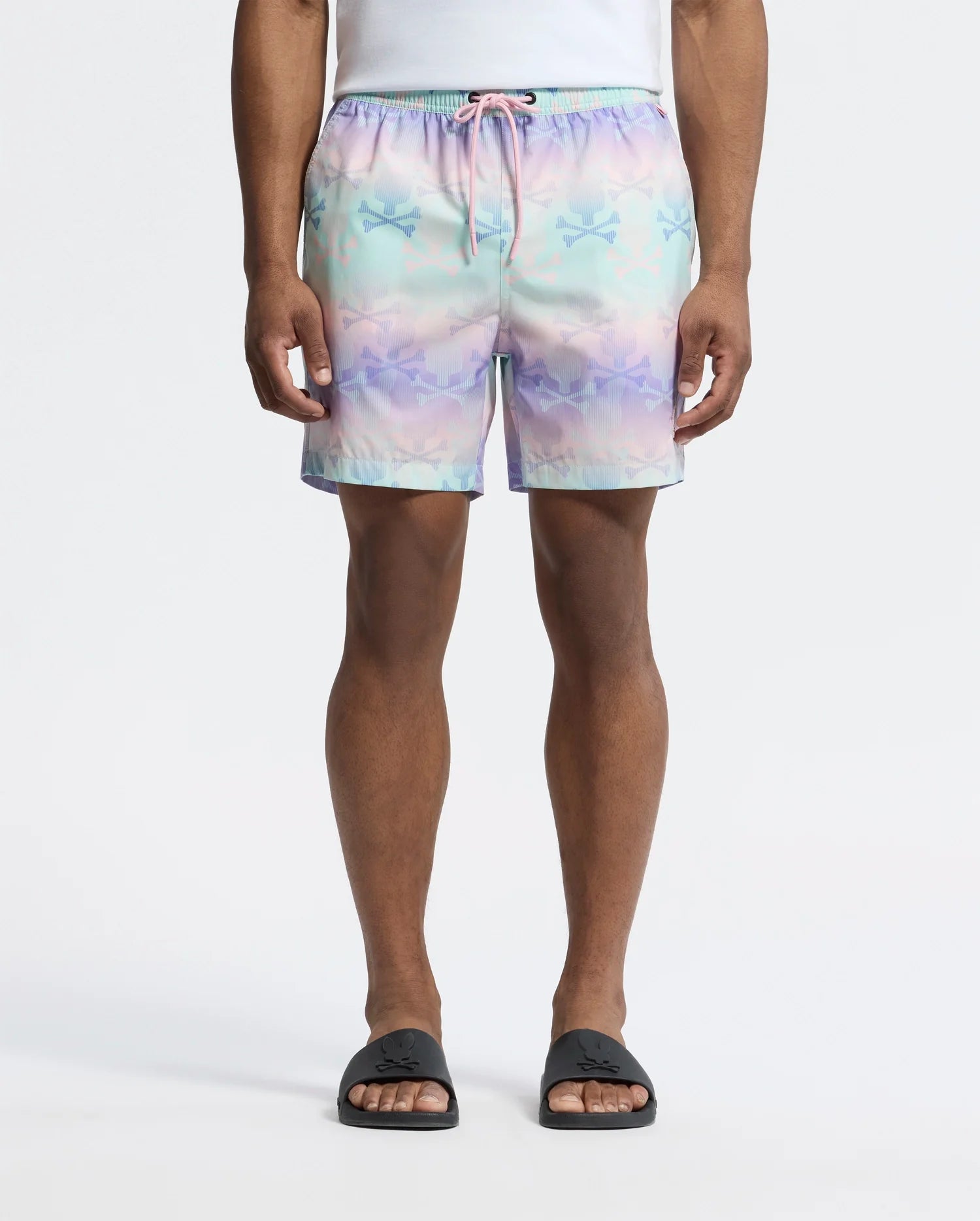 A person wears Psycho Bunny's MENS FORSTER BUNNY PRINTED SWIM TRUNK, a pastel-colored short with stars and bunnies featuring a pink drawstring, paired with a white shirt and black slide sandals against a plain white background. Their hands rest at their sides.