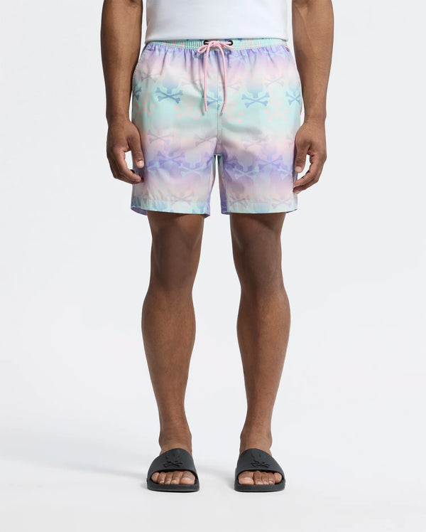 A person wears Psycho Bunny's MENS FORSTER BUNNY PRINTED SWIM TRUNK, a pastel-colored short with stars and bunnies featuring a pink drawstring, paired with a white shirt and black slide sandals against a plain white background. Their hands rest at their sides.
