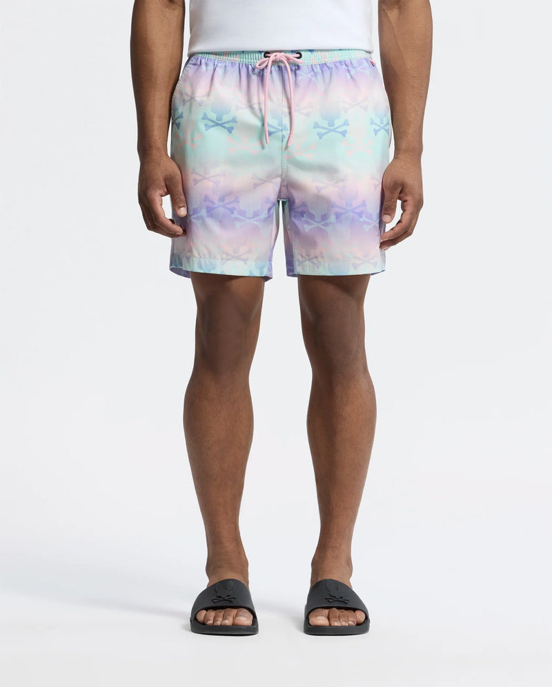 A person wears Psycho Bunny's MENS FORSTER BUNNY PRINTED SWIM TRUNK, a pastel-colored short with stars and bunnies featuring a pink drawstring, paired with a white shirt and black slide sandals against a plain white background. Their hands rest at their sides.