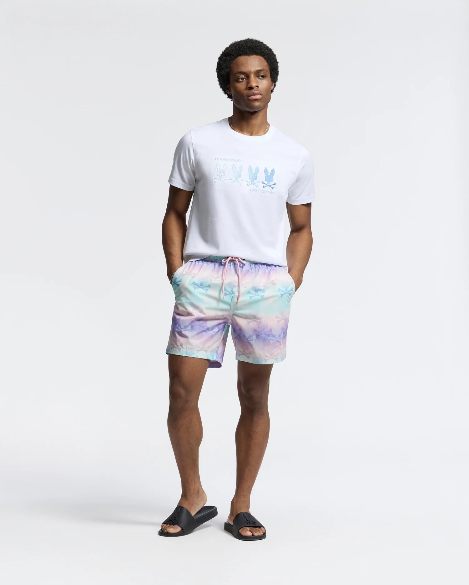 Wearing a Psycho Bunny MENS FORSTER BUNNY PRINTED SWIM TRUNK - B6W621F200, the man exudes effortless cool in pastel tie-dye shorts and black slide sandals, posing with hands in pockets against a white background.