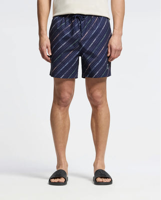 A modern man wears Psycho Bunny's MENS RENMARK PB PRINTED SWIM TRUNK, navy with a diagonal stripe design, a white t-shirt, and black slide sandals against a plain light-colored background. Perfect for style and function.
