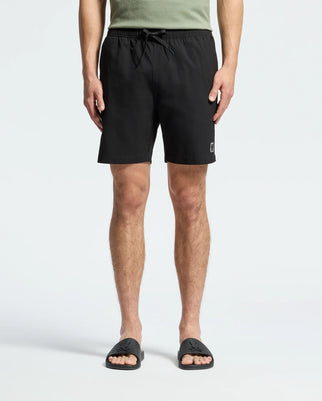 A person is wearing Psycho Bunny's MENS SAMOS 7 HYDROCHROMIC PRINTED SWIM TRUNK in black, a green T-shirt, and black slide sandals. The background is plain white, focusing on the sunsoaker style from the waist down.