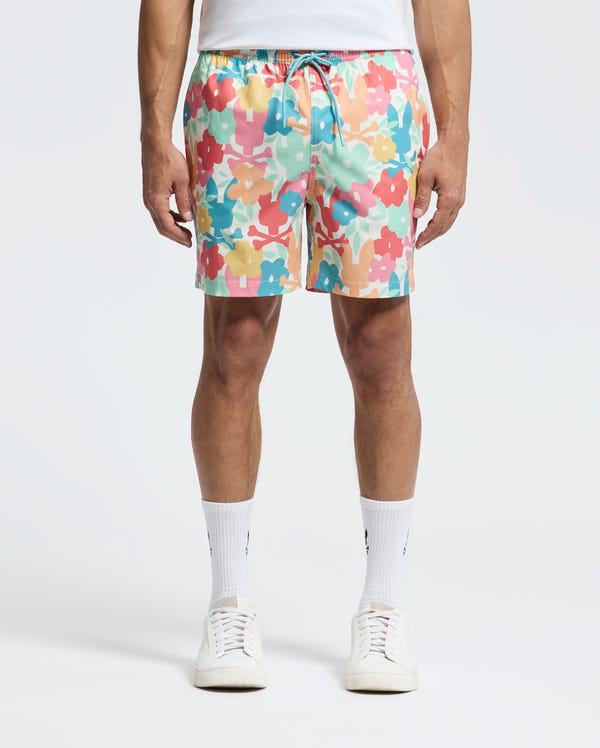 The person is wearing Psycho Bunny's vibrant MENS LEMMON ALL OVER PRINT SWIM TRUNK, blending comfort and style in pink, blue, yellow, and white. Paired with a crisp white shirt, socks, and sneakers against a light backdrop.