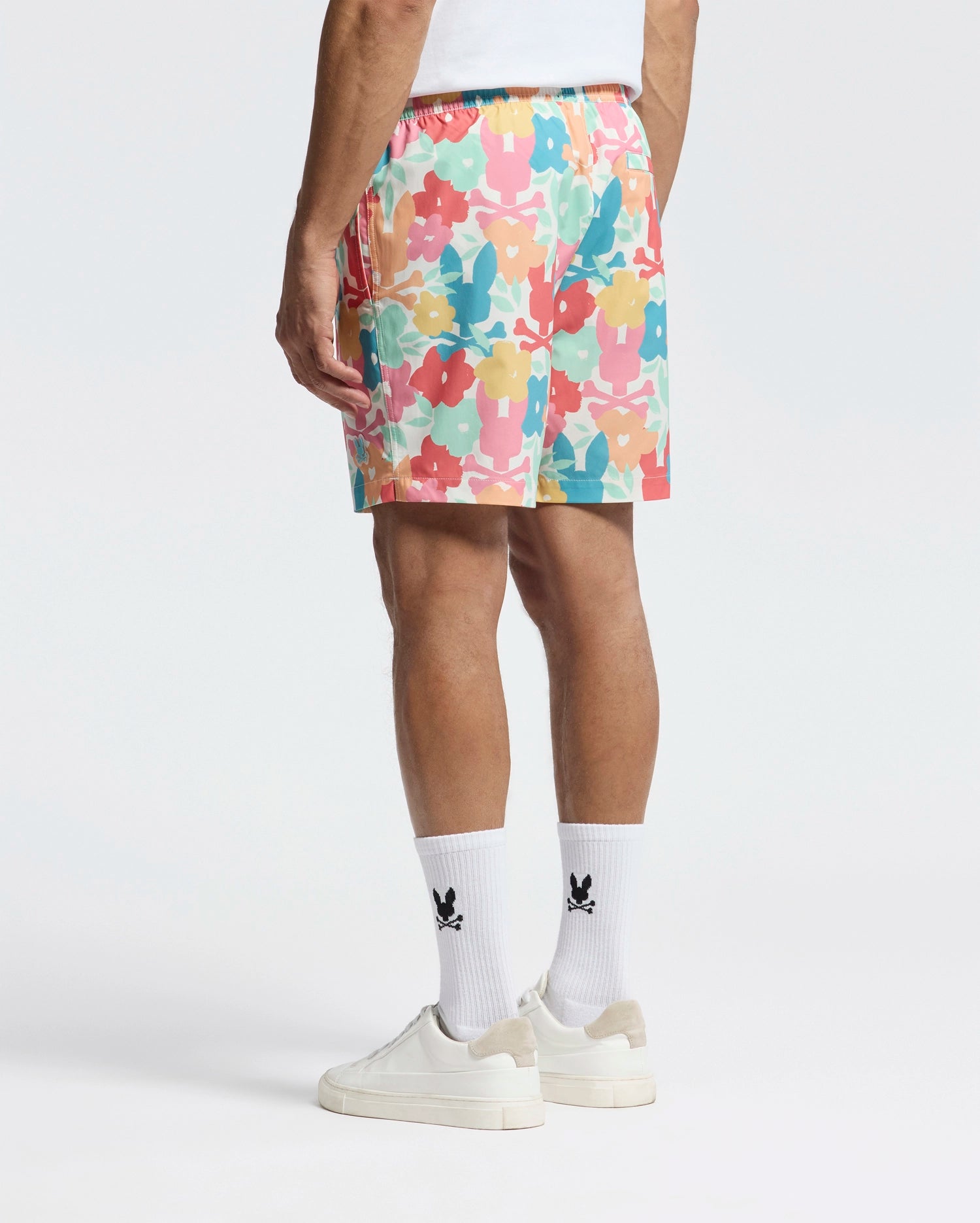 A person wearing Psycho Bunny's MENS LEMMON ALL OVER PRINT SWIM TRUNK with a white shirt and socks featuring black logos pairs them with white sneakers. The polyester mechanical stretch fabric offers comfort and style, all set against a plain white background.
