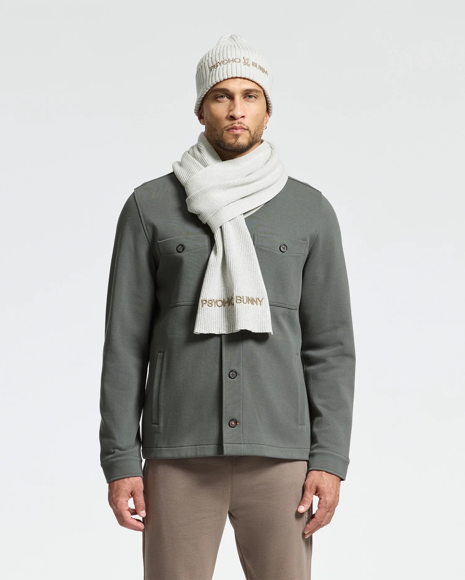 A man is wearing a green button-up jacket, beige pants, and the Psycho Bunny MENS BEANIE AND SCARF BOX SET - B6X353E200 in cream cotton with text detailing. He stands against a plain background, highlighting this cozy and stylish winter accessory set.