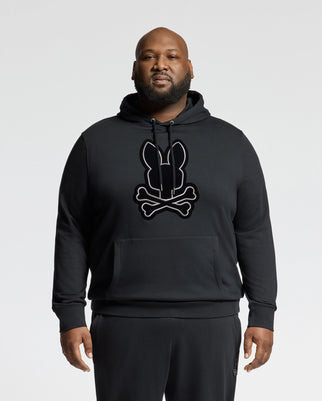 A person in a Psycho Bunny BIG AND TALL ALEXANDER HOODIE - B9H154E200, featuring a bunny and crossbones design, stands against a plain white background. This comfortable cotton blend pullover includes a front pocket and drawstrings. They are also wearing black pants.