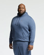 A man in the Psycho Bunny Mens Big and Tall Levy Essential Fleece Hoodie (B9H859D200) in blue stands facing the camera. This 100% cotton hoodie has a small chest logo and a convenient front pocket, shown against a plain, light background.