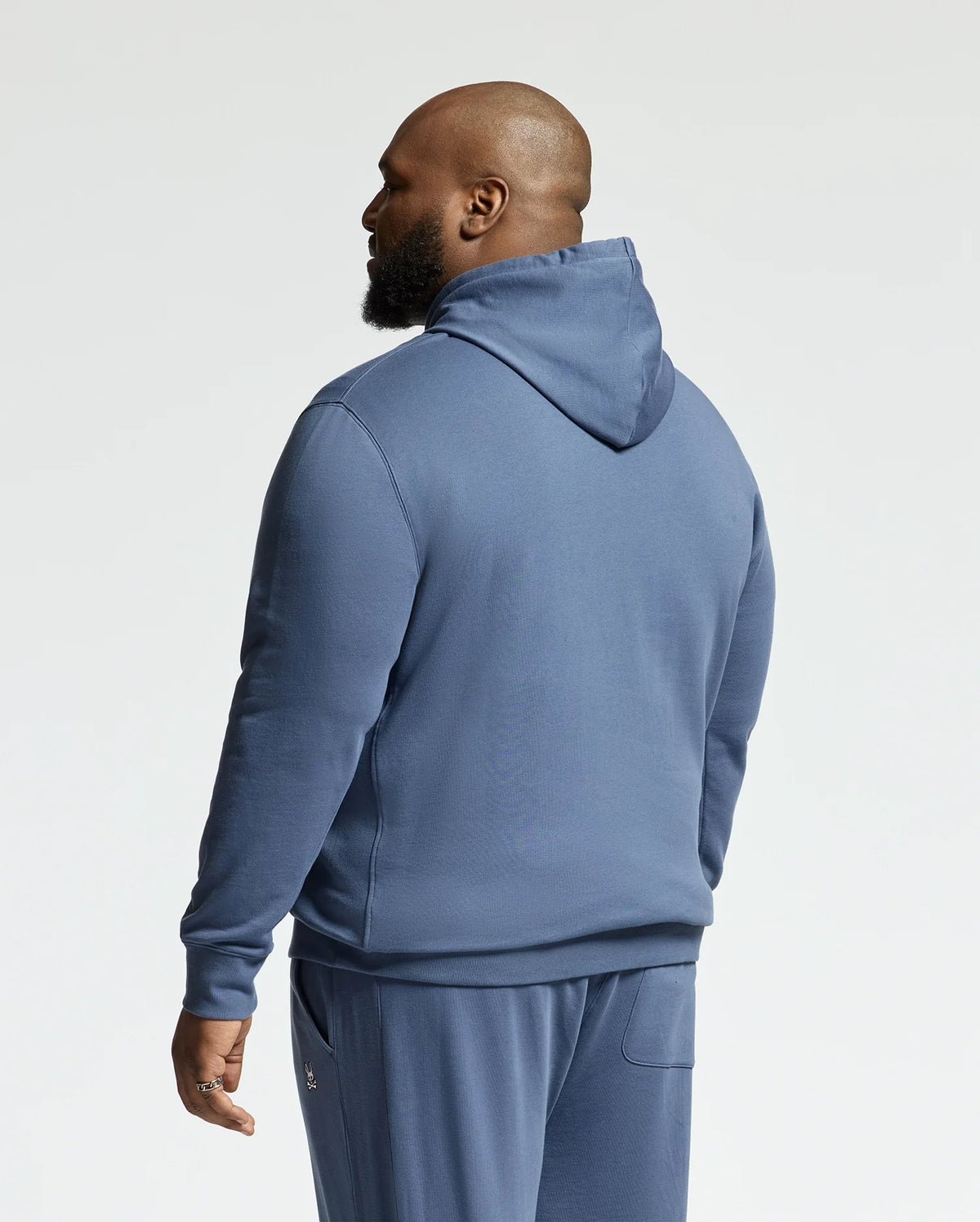 A man with a beard stands facing away, wearing the Psycho Bunny MENS BIG AND TALL LEVY ESSENTIAL FLEECE HOODIE (B9H859D200), styled against a plain light background.