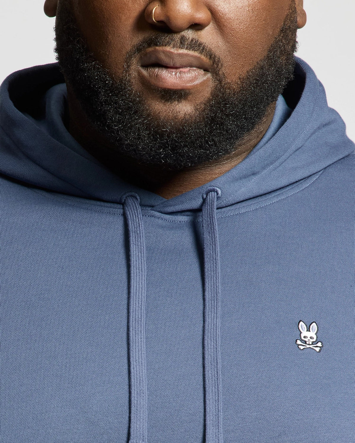 A close-up shows a person wearing the Psycho Bunny MENS BIG AND TALL LEVY ESSENTIAL FLEECE HOODIE, featuring a small white bunny and crossbones logo on the chest. The hoodie is blue, and part of the person's face and beard are visible.