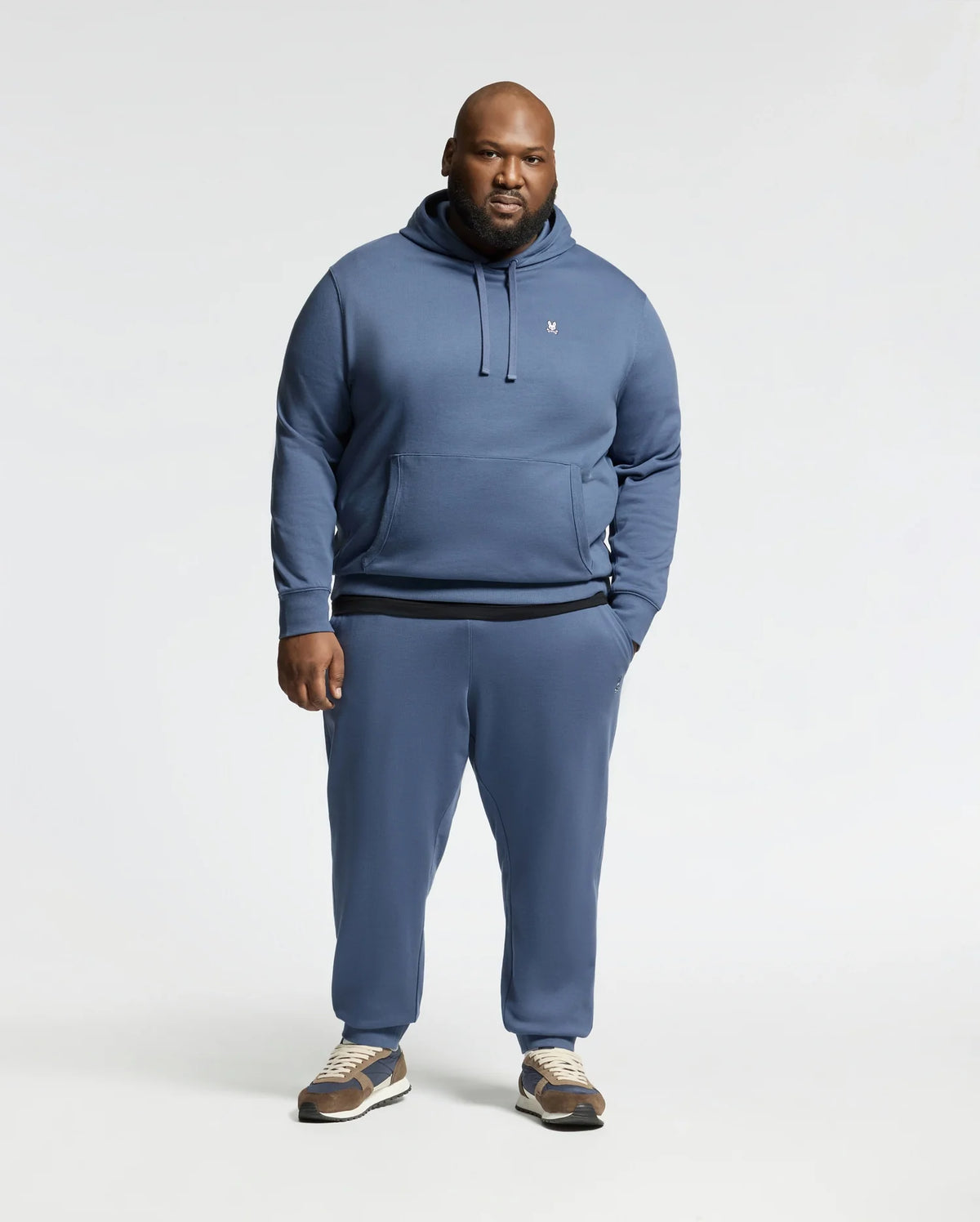 A man faces the camera wearing the Psycho Bunny MENS BIG AND TALL LEVY ESSENTIAL FLEECE HOODIE (B9H859D200) and matching sweatpants in blue, paired with brown and white sneakers against a plain, light-colored background.