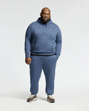 A man faces the camera wearing the Psycho Bunny MENS BIG AND TALL LEVY ESSENTIAL FLEECE HOODIE (B9H859D200) and matching sweatpants in blue, paired with brown and white sneakers against a plain, light-colored background.