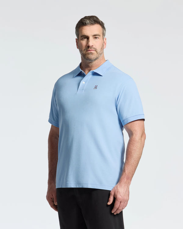 A man with short hair and a beard stands against a white background, wearing Psycho Bunny's BIG AND TALL CLASSIC PIQUE POLO - B9K001CRPC in light blue. His hands are relaxed at his sides, and the Pima Cotton shirt contrasts subtly with his dark pants.