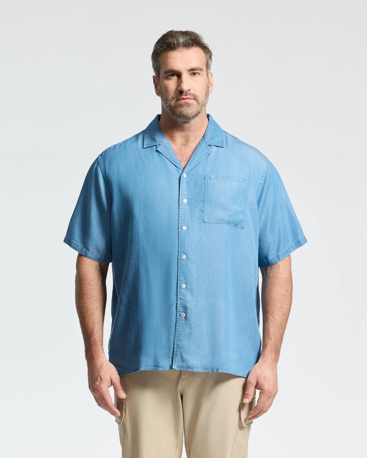 A man with short brown hair and a beard faces the camera against a plain background, wearing the Psycho Bunny MENS BIG AND TALL KERVIN LIGHTWEIGHT DENIM WOVEN SHIRT (B9Q467F200) in light blue and beige pants, giving a relaxed, casual vibe.