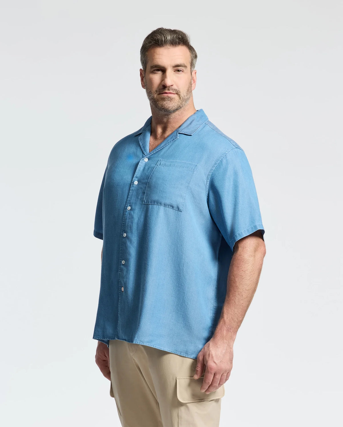 A man with short brown hair and a beard models the Psycho Bunny MENS BIG AND TALL KERVIN LIGHTWEIGHT DENIM WOVEN SHIRT (B9Q467F200) in light blue, styled casually with tan cargo pants against a plain background.