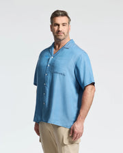 A man with short brown hair and a beard models the Psycho Bunny MENS BIG AND TALL KERVIN LIGHTWEIGHT DENIM WOVEN SHIRT (B9Q467F200) in light blue, styled casually with tan cargo pants against a plain background.