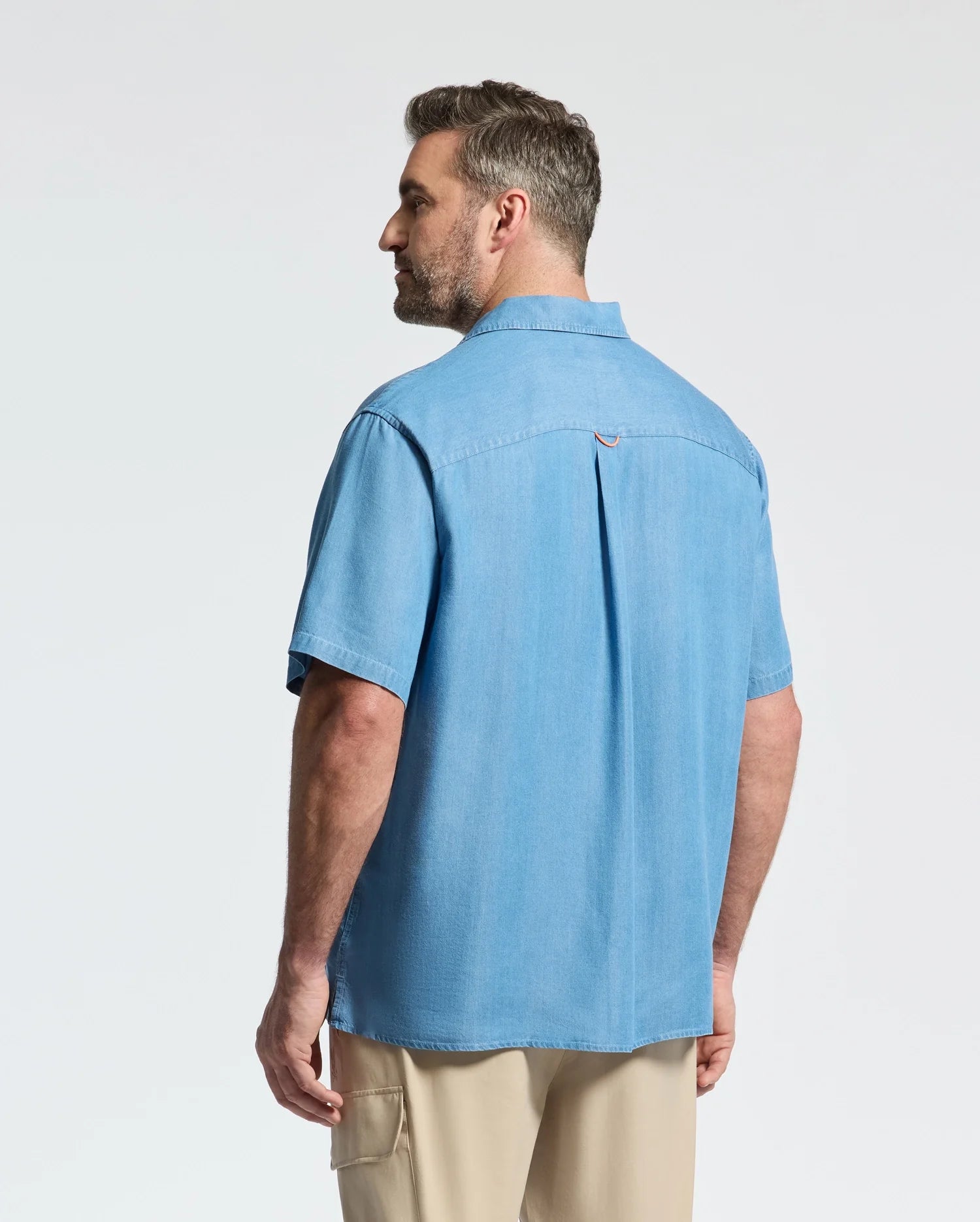 A man with short hair and a beard stands with his back to the camera in a Psycho Bunny MENS BIG AND TALL KERVIN LIGHTWEIGHT DENIM WOVEN SHIRT (B9Q467F200) and beige pants, creating a relaxed, casual vibe against a plain light background.